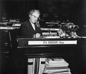 A woman in glasses writes at a desk with a placard on the front that says Eva Anderson Chelan. 